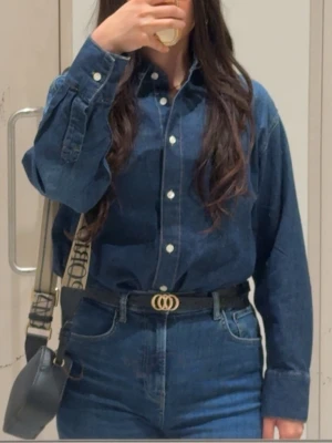 Blå jeansskjorta med knappar - Klassisk blå jeansskjorta med vita knappar och lång ärm. Skjortan har en avslappnad passform och är tillverkad i ett robust bomullsmaterial. Perfekt att styla med jeans för en cool denim-on-denim look.