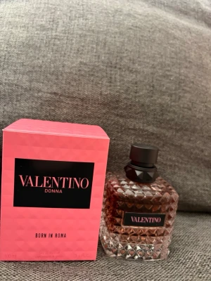 Valentino Donna Born In Roma parfym - Valentino Donna Born In Roma parfym med en lyxig, rosa kartong och en elegant glasflaska med nitar och svart lock. Flaskan har en modern, stilren design och etikett med Valentino-loggan. Perfekt för dig som gillar exklusiva dofter och snygg inredningsdetalj.