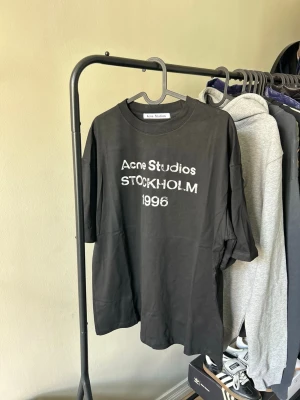 Svart Acne Studios t-shirt - Säljer en svart t-shirt från Acne Studios med tryck i vitt framtill där det står 'Acne Studios STOCKHOLM 1996'. T-shirten har rund hals och korta ärmar. Perfekt för en avslappnad och trendig stil.