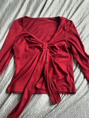 Vinröd långärmad topp med knyt - Snygg vinröd långärmad topp med knyt framtill som ger en cool omlottkänsla. Toppen har en djupare ringning och är gjord i ett mjukt, stretchigt material som känns skönt mot huden. Perfekt för dig som vill ha en trendig och bekväm look.
