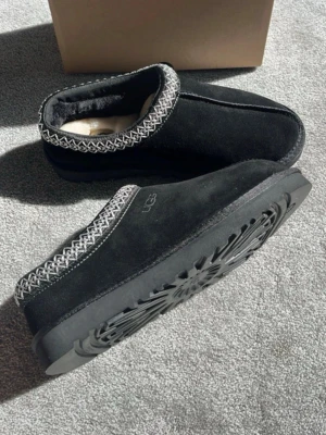 Svarta UGG tofflor med mönstrad kant - Storleken är lite konstig dem är ganska små i storlek skulle säga att dem är 41 inte 41             Säljer ett par svarta UGG tofflor i mocka med mjuk, fluffig insida och en snygg gråvit mönstrad kant runt öppningen. Yttersulan är robust och har ett tydligt mönster för bra grepp. Perfekta för att hålla fötterna varma hemma.