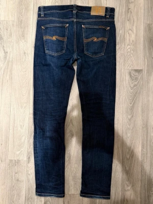 Nudie Jeans Lean Dean Dry Mörkblå Jeans W32/L32 - Slim Tapered Fit - Nudie Jeans Lean Dean ”Dry 16 Dips” mörkblå Dry-jeans med slim tapered-passform, normalhög midja och avsmalnande ben, storlek W32/L32, midja 41,5 cm, ytterbenslängd 101,5 cm, benöppning 16 cm, modellen är 185cm & väger 65kg, i fint skick.
