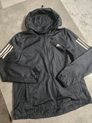 Svart vindjacka från Adidas - Svart vindjacka från Adidas med huva och dragkedja framtill. Jackan har vita klassiska Adidas-ränder på ärmarna och logga på bröstet. Tillverkad i lätt och slitstarkt material, perfekt för blåsiga dagar. Två fickor framtill och elastiska muddar vid ärmslut.
