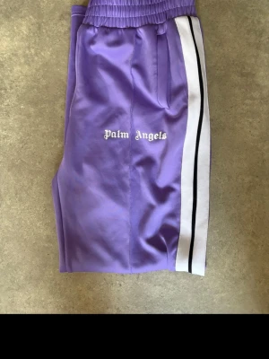 Lila shorts från Palm Angels - Snygga lila shorts från Palm Angels med vita och svarta ränder längs sidorna. Shortsen har elastisk midja och fickor, samt logga broderad framtill. Perfekta för en avslappnad och trendig look.