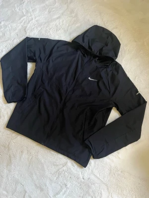 Svart vindjacka med huva från Nike - Snygg svart vindjacka från Nike med huva och dragkedja framtill. Jackan har två sidofickor och en diskret Nike-logga på bröstet. Tillverkad i lätt och vindtätt material, perfekt för blåsiga dagar. Enkel och stilren design som passar till det mesta.