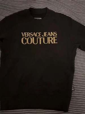 Svart sweatshirt Versace Jeans Couture - Snygg svart sweatshirt från Versace Jeans Couture med stor guldfärgad logga framtill som ja använt en gång o de va bara test. Klassisk rund halsringning och ribbade muddar vid ärmslut och nederkant. Perfekt för dig som gillar streetwear och vill ha en statementtröja.
