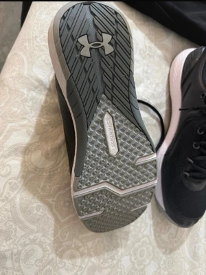 Svarta Under Armour sneakers - Snygga svarta sneakers från Under Armour med vit sula och logga på sidan. Ovandelen är i mesh för bra andningsförmåga och skon har snörning. Sulan har Charged Cushioning för extra komfort och grepp. Perfekta för dig som gillar sportig stil.