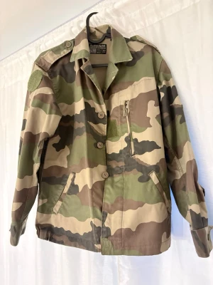 Klassisk camouflage vindjacka - Säljer en grön och beige camouflage vindjacka med knappar framtill och flera fickor. Jackan har lång ärm, axelklaffar och är tillverkad i bomull. Perfekt för dig som gillar streetstyle eller militärinspirerad look.