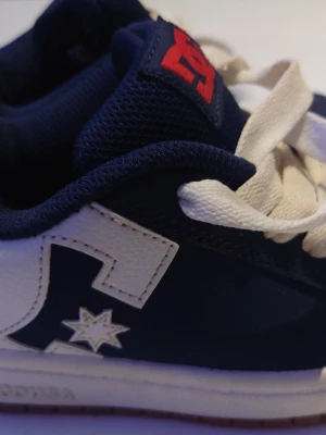 Marinblå och vita DC sneakers - Snygga sneakers från DC i marinblått och vitt med tydlig DC-logga på sidan och röd detalj på plösen. Skorna har vita skosnören och en stjärndetalj på sidan. Ovandelen är i mocka och läder för en cool kontrast.