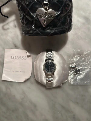Klocka från Guess - Stilren klocka från Guess med silverfärgad länkarmband och svart urtavla. Klockan har klassisk rund form och detaljerade markeringar på urtavlan. Levereras med extra länkar, originalpåse och garantiinformation (garantin har gått ut dock). Kockan behöver nya batterier.