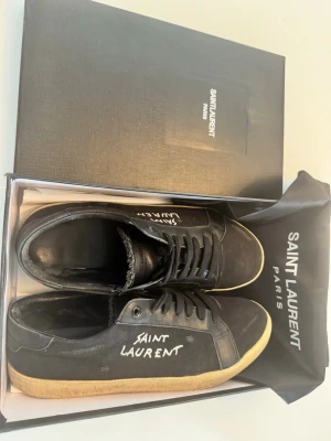 Saint Laurent svarta sneakers - Svarta sneakers från Saint Laurent med vit logga på sidan och beige sula. Skorna har en klassisk låg profil, snörning och är tillverkade i en mix av läder och mocka. Perfekta för dig som gillar stilrena och exklusiva sneakers.