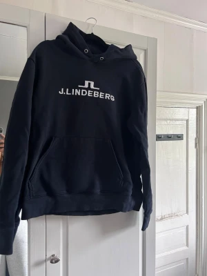 Marinblå hoodie från J.Lindeberg - Snygg marinblå hoodie från J.Lindeberg med stor vit logga och text på bröstet. Ficka framtill.