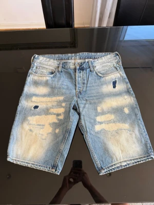 H&M jeansshorts - Säljer ett par ljusblå jeansshorts med slitna detaljer och lagningar framtill. Klassisk femficksmodell med normal passform och bälteshällor. Perfekta för en avslappnad och trendig look i sommar.