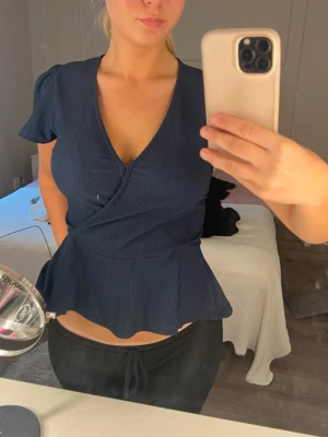 Mörkblå omlottopp med volang - Snygg mörkblå omlottopp med v-ringning och volang nedtill. Toppen har korta ärmar och markerad midja som ger en feminin siluett. Perfekt att styla med både jeans och kjol för en trendig look.