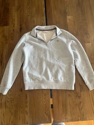 Zip hoodie - Ljusgrå sweatshirt med halv dragkedja och ståkrage. Tröjan har ribbade muddar vid ärmslut och nederkant samt diskret broderad logga på bröstet. Insidan av kragen har randiga detaljer. Perfekt för en avslappnad och stilren look.