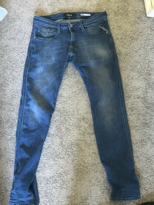 Blå slim fit jeans från Replay - Säljer ett par blå slim fit jeans från Replay med klassisk femficksdesign och snygga slitningar framtill. Jeansen har dragkedja och knappstängning samt detaljerade bakfickor med Replay-logga. Materialet är mjukt denim som sitter skönt. W33 L34 men midjan passar som 32-33 och längden 32-33