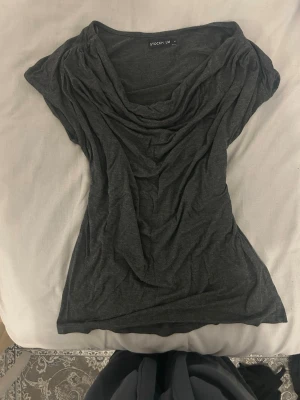 Mörkgrå draperad topp y2k 2000s - Snygg mörkgrå topp med draperad halsringning och korta ärmar. Toppen har en loose passform och är tillverkad i ett mjukt, tunt material som känns skönt mot huden. Perfekt att styla med jeans eller kjol för en avslappnad look.
