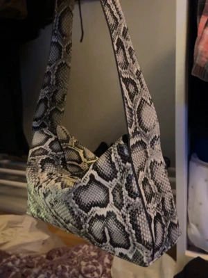 Axelremsväska i ormmönstrat konstläder - Säljer en trendig axelremsväska med svartvitt ormmönster. Väskan har en bred axelrem och boxig form, perfekt för att ge din outfit en edgy touch. Materialet är konstläder med tydlig struktur och mönster som liknar ormskinn.