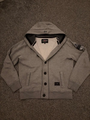 Grå cardigan hoodie med patch - Tjenare! Säljer en fet speciell grå hoodie med knappar o en patch den är S men är lite stor i bredden den sitter rätt bra på mig i längd som är 185.