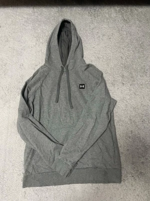 Grå hoodie från Under Armour - Snygg grå hoodie från Under Armour med klassisk logga på bröstet. Hoodien har dragsko i huvan och ribbade muddar vid ärmslut och nederkant. Tillverkad i mjukt material som passar perfekt till chill eller träning.