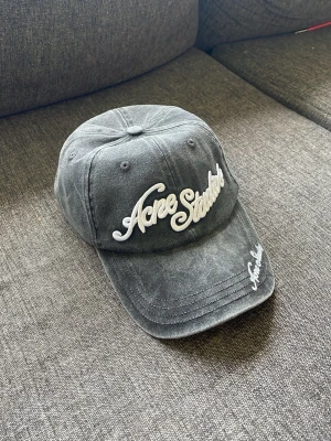  Acne Studios keps - Säljer en grå keps från Acne Studios med broderad vit logga framtill och på skärmen. Kepsen har klassisk böjd skärm och justerbar rem bak. Tillverkad i tvättad bomull för en avslappnad look.