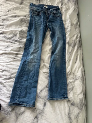 Blå bootcut jeans med slitningar - Säljer ett par blå bootcut jeans med snygga slitningar och klassiska fem fickor. Jeansen har en lätt tvättad look och raka ben som går ut lite nedtill. Perfekta för dig som gillar en avslappnad men trendig stil.