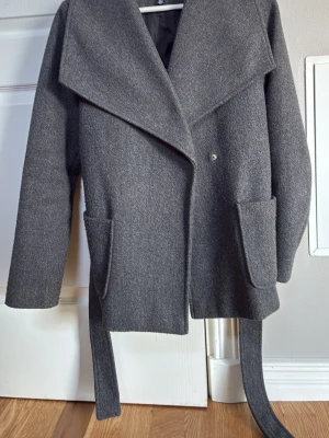 Grå kappa med bred krage och skärp - Stilren grå kappa med bred krage och knytbart skärp i midjan. Kappan har två stora framfickor och en avslappnad passform. Perfekt för kyliga dagar och enkel att matcha med olika outfits.