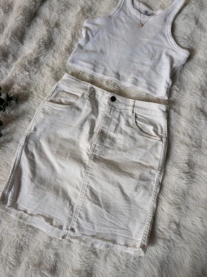 Vit jeanskjol med rå kant - Säljer en vit jeanskjol i kort modell med rå, fransig kant nedtill. Kjolen har klassiska fem fickor, bälteshällor och knappstängning framtill. Perfekt till sommaren för en avslappnad och trendig look.