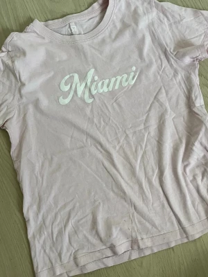 Ljusrosa t-shirt med Miami-tryck - Säljer en ljusrosa t-shirt med vit text 'Miami' på bröstet. T-shirten har rund halsringning och korta ärmar. Perfekt för en avslappnad och trendig look.