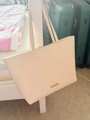 Stor beige tote handväska - Snygg och stilren beige tote handväska i slätt konstläder med dubbla axelband. Väskan har en enkel design med en liten metallbricka framtill. Perfekt för dig som vill ha plats för allt du behöver under dagen.