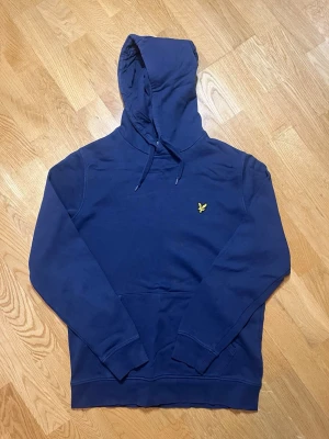 Mörkblå hoodie Lyle & Scott - Säljer denna snygga mörkblåa hoodie från Lyle & Scott med klassisk gul logga på bröstet. Hoodien har dragsko i huvan och en stor magficka. Tillverkad i mjuk bomull för skön känsla och avslappnad stil. Perfekt för dig som gillar enkel och stilren design. OBS! Det finns ett litet hål på vänstra axeln av hoodien. Hålet syns däremot inte på långt håll men går att sy ihop. Vid frågor, extra bilder eller liknande är det bara att kontakta mig!