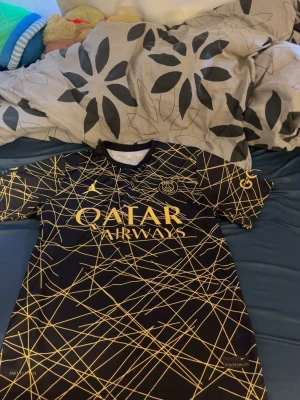 PSG Neymar Jr matchtröja svart/guld - Snygg PSG matchtröja i svart med guldiga detaljer och grafiskt mönster. Framsidan har Qatar Airways-logga och PSG-emblem, baksidan har Neymar Jr och nummer 10 i guld. Tillverkad i lätt och ventilerande material, perfekt för fotboll eller träning.
