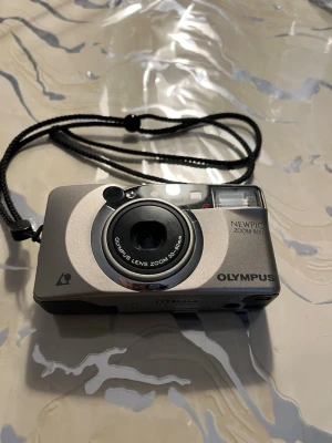 Olympus Newpic Zoom 600 - APS kompaktkamera - Snygg kompakt digitalkamera från Olympus, modell Newpic Zoom 600. Kameran är silverfärgad med svarta detaljer och har ett inbyggt zoomobjektiv (30-60mm). Praktisk handledsrem i svart medföljer. Perfekt för dig som vill ha en smidig kamera att ta med överallt.