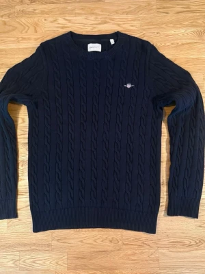 Mörkblå stickad GANT tröja - Kabelstickad mörkblå tröja från GANT med rund halsringning och ribbade muddar. Tröjan har ett broderat GANT-emblem på bröstet och är tillverkad i mjukt material som passar perfekt till höst och vinter. Klassisk och tidlös design.