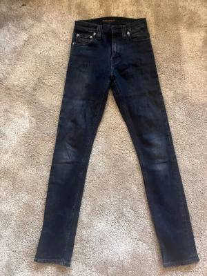 Nuddie jeans - Säljer ett par mörkblå skinny jeans från Nudie Jeans med klassisk femficksdesign och hög midja. Jeansen är tillverkade i stretchigt denimtyg som sitter tight hela vägen ner. Perfekta för dig som gillar en smal siluett och tidlös look.