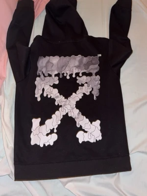 Svart Off-White hoodie med tryck - Svart hoodie från Off-White med stor grafisk logga i grått och vitt på ryggen och mindre tryck framtill. Klassisk huva med dragsko och känguruficka. Perfekt för dig som gillar streetwear och vill sticka ut med en unik design.