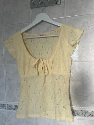 Gul spetsblus med rosett och volangärm - Söt gul blus i spets med korta volangärmar och knyt framtill. Blusen har ett broderat blommönster med en super fin U-ringning. Toppen går att både klä upp och klä ner med sin fina gula färg💛