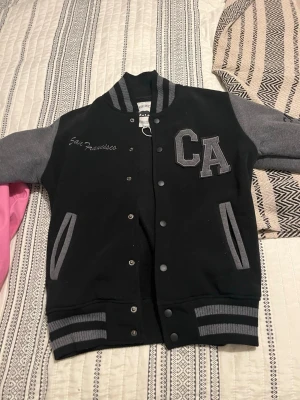 Svart och grå collegejacka CA - Snygg svart och grå collegejacka från California Citywear med broderad text 'San Francisco' och stora bokstäver 'CA' på bröstet. Jackan har grå ärmar, randiga muddar och knappar framtill. Perfekt för en sportig och avslappnad stil.