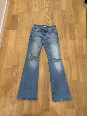 Ljusblå bootcut jeans med hål - Säljer ett par ljusblå bootcut jeans med slitningar och stora hål på båda knäna. Jeansen har dubbla knappar i midjan, klassiska fem fickor och är tillverkade i denim. Perfekta för en avslappnad och trendig look. Väldigt trendinga eftertraktade fårn Nelly. Pris kan diskuteras vid snabb affär 