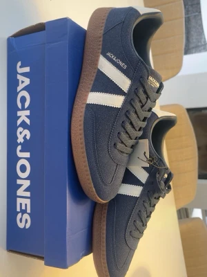 Blå sneakers från Jack & Jones - Snygga blå sneakers från Jack & Jones med vita detaljer på sidan och klassisk gummisula. Ovandelen är i mockaliknande material och skorna har låga skaft. Perfekta för en stilren och avslappnad look. Dessa skorna är helt nya med lappen på