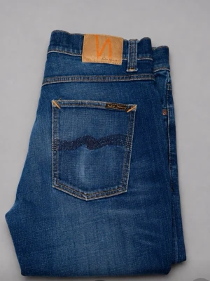  Nudie Jeans slitningar  - Snygga blå jeans från Nudie Jeans med klassisk femficksmodell. Jeansen har kontrastsömmar, läderpatch med logga bak och vågig detalj på bakfickorna. Tillverkade i slitstark denim, perfekta för en avslappnad stil.