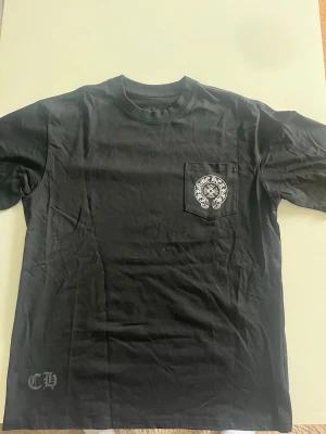 Svart Chrome Hearts t-shirt - Snygg svart t-shirt från Chrome Hearts med vit logga på bröstfickan och tryck på ärmen. Klassisk passform och rund hals.