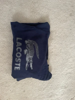Mörkblå hoodie från Lacoste - Snygg mörkblå hoodie från Lacoste med det klassiska krokodillogot i grått broderat på bröstet. Hoodien har långa armar  och en stor luva. Perfekt för dig som gillar stilrena och sportiga plagg med tydlig logga.