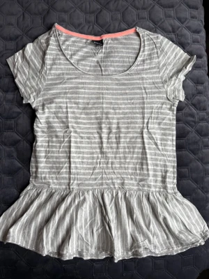 Randig peplumtopp från Gina Tricot - Säljer en ljusgrå och vit randig peplumtopp från Gina Tricot. Toppen har korta ärmar, rund halsringning och volang nedtill. Perfekt för dig som gillar en avslappnad men söt stil.