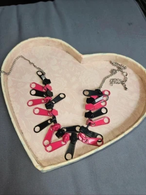 Halsband med svarta och rosa dragkedjor - Säljer ett kreativt halsband gjort av svarta och rosa dragkedjor på en silverfärgad kedja. Dragkedjorna hänger tätt i ett mönster som ger ett edgy och alternativt uttryck. Perfekt accessoar för dig som gillar statement-smycken och vill sticka ut.  Kan även hämtas I Partille  J
