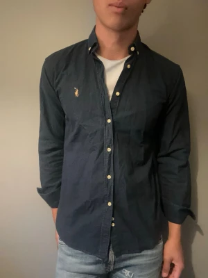 Mörkblå skjorta från Ralph Lauren - Snygg mörkblå skjorta från Ralph Lauren med klassisk krage och knappar framtill. Skjortan har lång ärm och den ikoniska broderade loggan på bröstet. Perfekt för en stilren look.
