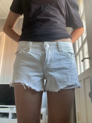 Vita jeansshorts med fransig kant - Säljer ett par vita jeansshorts med fransig kant och råa avslut. Shortsen har fickor både fram och bak, samt bälteshällor och knappstängning. Perfekta för varma dagar och ger en avslappnad look.