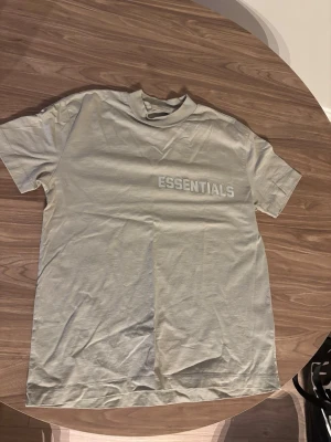 Beige FEAR OF GOD Essentials t-shirt - Snygg beige t-shirt från Essentials med tryckt logga på bröstet. T-shirten har en avslappnad passform, rund halsringning och är tillverkad i mjukt bomullsmaterial. Perfekt för en clean och stilren look.