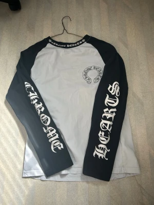 Långärmad Chrome Hearts t-shirt - Säljer en långärmad t-shirt från Chrome Hearts med svartvita kontraster. Tröjan har svarta ärmar och vit kropp, samt stora Chrome Hearts-tryck på ärmarna och ryggen. Rund halsringning med logga och tillverkad i mjuk bomull.