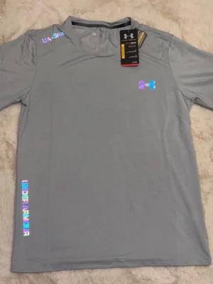 Grå Under Armour t-shirt - Snygg grå t-shirt från Under Armour med holografiska loggor på bröstet och vid nederkanten. T-shirten har korta ärmar och är tillverkad i ett lätt och funktionellt material som passar perfekt till träning.